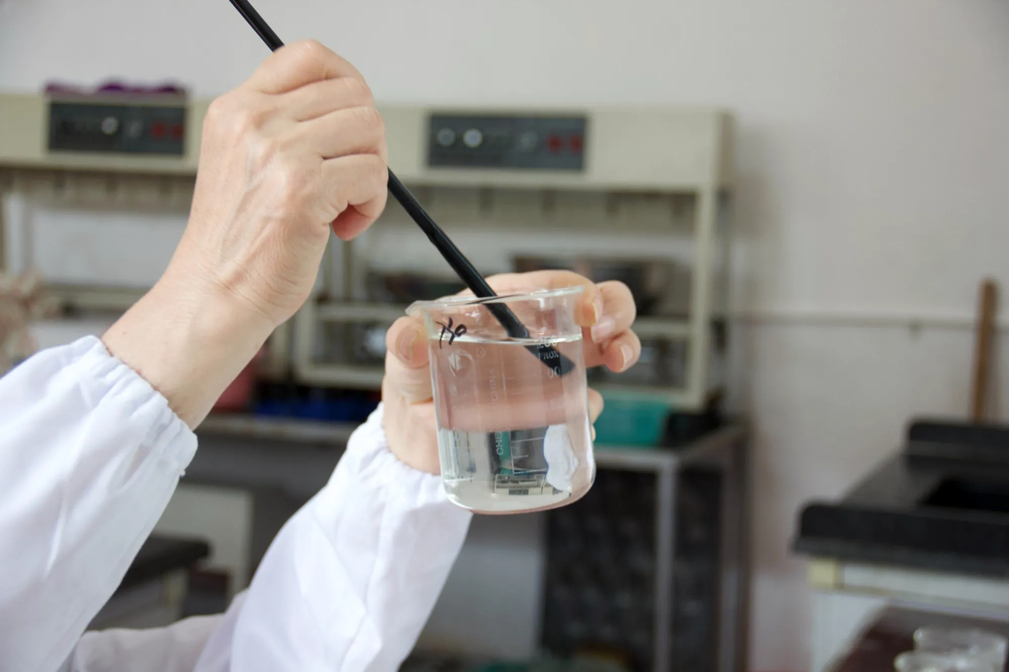 Técnico del laboratorio de Sentai agitando una solución de CMC transparente y viscosa en un vaso de precipitados de vidrio con una varilla de vidrio